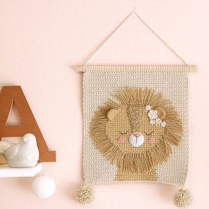 May include: A beige crocheted wall hanging with a lion face in the center. The lion has a brown mane, closed eyes, a pink nose, and pink flowers on its head. The wall hanging has two beige pom-poms at the bottom.