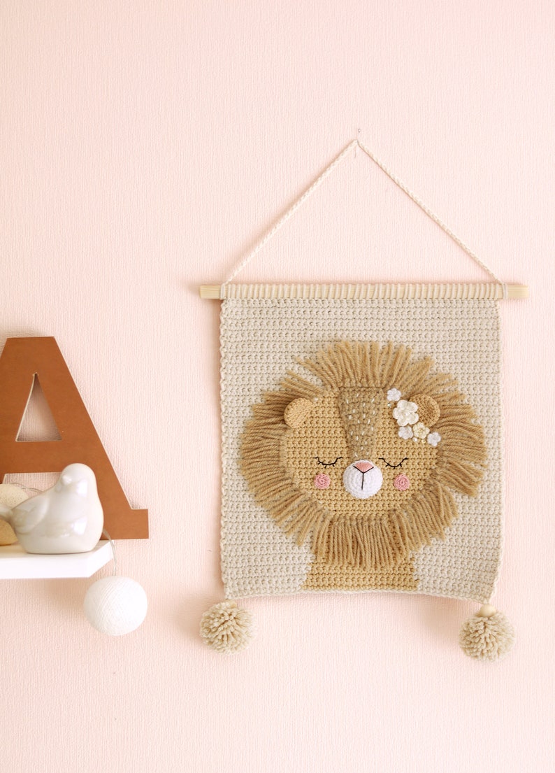 May include: A beige crocheted wall hanging with a lion face in the center. The lion has a brown mane, closed eyes, a pink nose, and pink flowers on its head. The wall hanging has two beige pom-poms at the bottom.