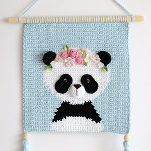 May include: A crocheted wall hanging featuring a panda with a flower crown. The panda is white with black ears, eyes, nose, and markings. The background is light blue and the hanging has two light blue tassels.