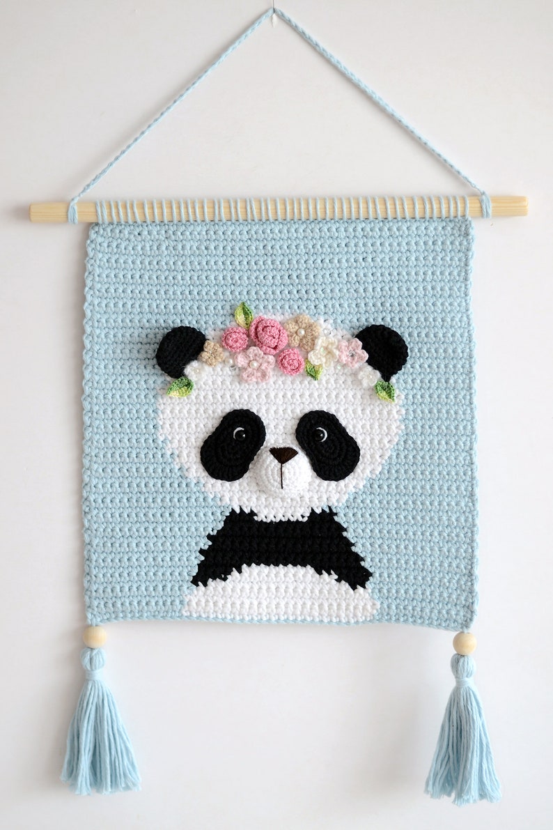May include: A crocheted wall hanging featuring a panda with a flower crown. The panda is white with black ears, eyes, nose, and markings. The background is light blue and the hanging has two light blue tassels.