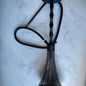 May include: A black hookah with a transparent, pear-shaped base and a dark stem. The stem has a bamboo-like design. The hose and top components are also black. The word "LADANA" is printed vertically on the base.