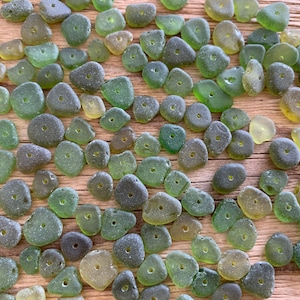 Puede incluir: Un conjunto de cuentas de vidrio marino en tonos verdes y verde-amarillo, cada una con un peque&ntilde;o agujero en el centro. Las cuentas, con sus formas irregulares y texturas esmeriladas, est&aacute;n dispuestas sobre una superficie de madera.