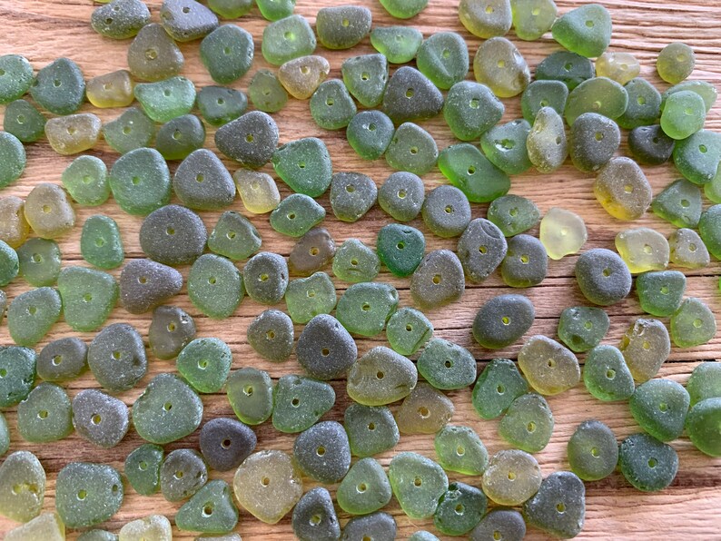 Puede incluir: Un conjunto de cuentas de vidrio marino en tonos verdes y verde-amarillo, cada una con un peque&ntilde;o agujero en el centro. Las cuentas, con sus formas irregulares y texturas esmeriladas, est&aacute;n dispuestas sobre una superficie de madera.
