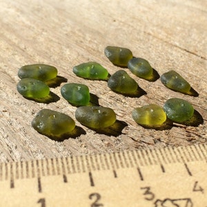 Puede incluir: Una colecci&oacute;n de piezas de vidrio de mar verde oliva, cada una con una forma suave y redondeada, dispersas sobre una superficie de madera envejecida. Una regla es visible en primer plano, proporcionando escala. El vidrio tiene una cualidad transl&uacute;cida.
