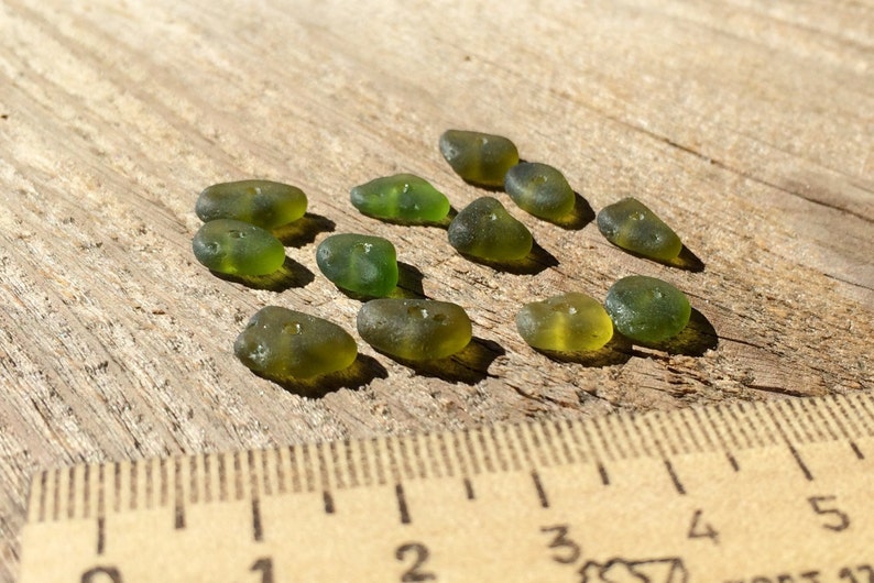 Puede incluir: Una colecci&oacute;n de piezas de vidrio de mar verde oliva, cada una con una forma suave y redondeada, dispersas sobre una superficie de madera envejecida. Una regla es visible en primer plano, proporcionando escala. El vidrio tiene una cualidad transl&uacute;cida.
