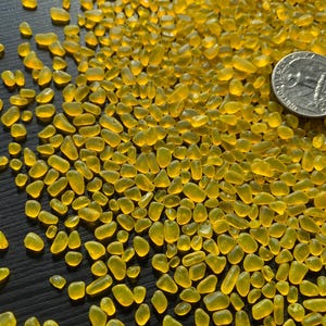 May include: A close-up shot of numerous small, translucent, yellow glass beads. A British coin is visible among the beads, providing a size reference. The beads are scattered on a dark surface.
