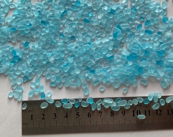 May include: A close-up of a pile of small, light blue glass pebbles. The pebbles are scattered on a white surface and a ruler is visible at the bottom of the image. The ruler is marked in millimeters and shows measurements from 0.5 to 14.