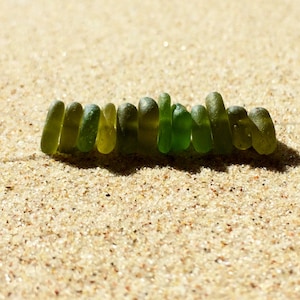 Puede incluir: Un collar hecho de cuentas de vidrio marino verde, sobre arena. Las cuentas son de diferentes tonos de verde, de forma oblonga y transl&uacute;cidas. Est&aacute;n ensartadas en un cord&oacute;n fino, casi invisible. La arena es de color marr&oacute;n claro y texturizada.