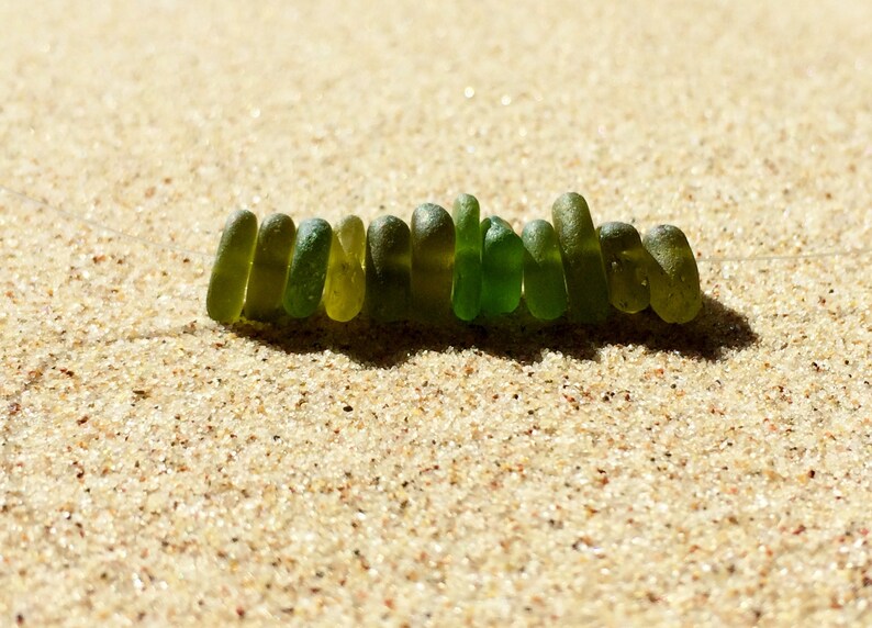 Puede incluir: Un collar hecho de cuentas de vidrio marino verde, sobre arena. Las cuentas son de diferentes tonos de verde, de forma oblonga y transl&uacute;cidas. Est&aacute;n ensartadas en un cord&oacute;n fino, casi invisible. La arena es de color marr&oacute;n claro y texturizada.