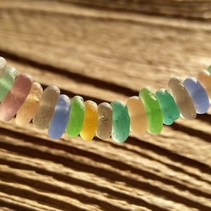 Könnte beinhalten: Eine bunte Halskette aus glatten, abgerundeten Meerglassperlen in Blautönen, Grün, Gelb und Rosa. Die Perlen sind auf einem dünnen Draht aufgefädelt und in einem versetzten Muster angeordnet.