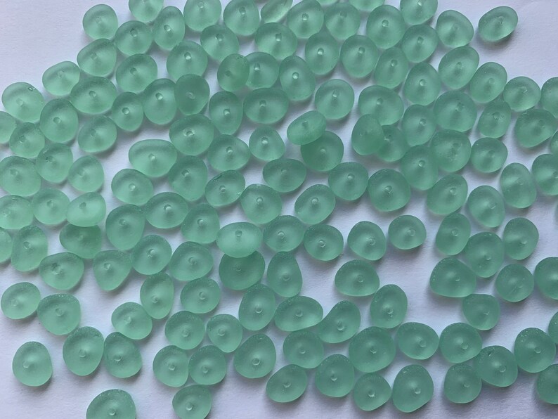 Op de afbeelding: Een verzameling lichtgroene glazen kralen met een matte afwerking. De kralen zijn gevormd als kleine, onregelmatige kiezelsteentjes met een gat in het midden.
