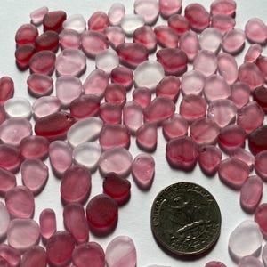 May include: A collection of smooth, tumbled sea glass in shades of pink and red, with a US quarter for size comparison. The glass pieces vary in shape and opacity, ranging from translucent to opaque.