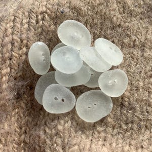 Puede incluir: Una colecci&oacute;n de botones de vidrio de mar blanco liso con dos agujeros cada uno. Los botones est&aacute;n dispuestos en una pila sobre una superficie de punto marr&oacute;n.