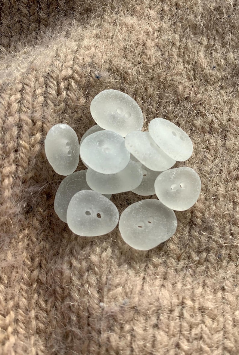 Puede incluir: Una colecci&oacute;n de botones de vidrio de mar blanco liso con dos agujeros cada uno. Los botones est&aacute;n dispuestos en una pila sobre una superficie de punto marr&oacute;n.