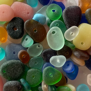 Puede incluir: Una colección de cuentas de vidrio de mar de colores con agujeros perforados en el centro. Las cuentas son de varias formas y tamaños, incluyendo redondas, ovaladas y en forma de lágrima. Los colores incluyen azul, verde, amarillo, rosa y marrón.