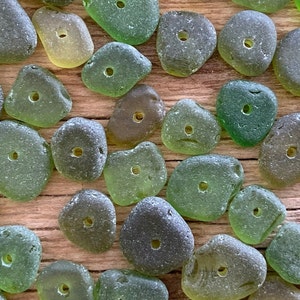 Puede incluir: Una colecci&oacute;n de cuentas de vidrio marino en varios tonos de verde y gris, cada una con un agujero perforado. Las cuentas est&aacute;n dispuestas sobre una superficie de madera, mostrando sus formas lisas y redondeadas y sus variaciones naturales de color y textura. Ideal para la creaci&oacute;n de joyas.