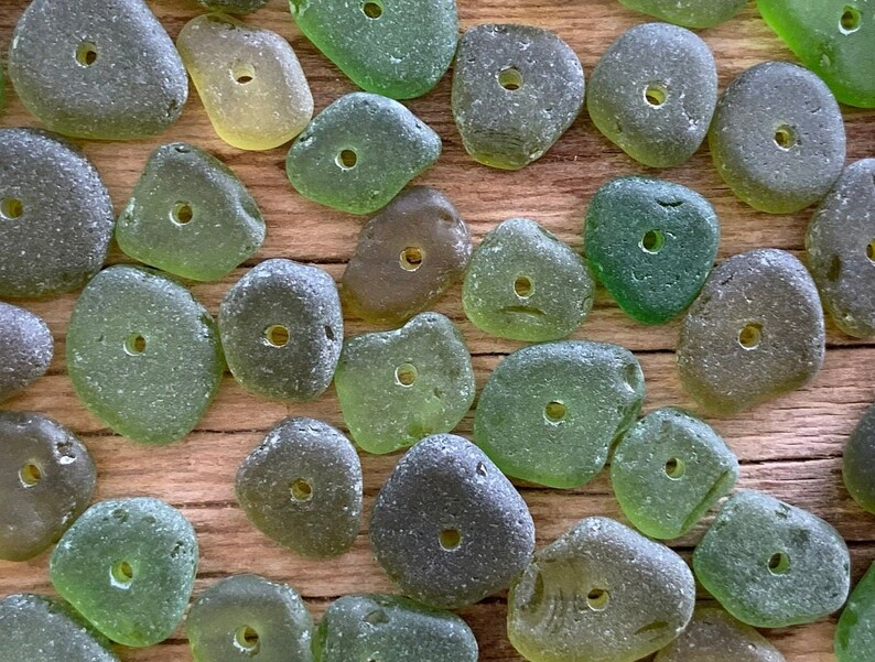 Puede incluir: Una colecci&oacute;n de cuentas de vidrio marino en varios tonos de verde y gris, cada una con un agujero perforado. Las cuentas est&aacute;n dispuestas sobre una superficie de madera, mostrando sus formas lisas y redondeadas y sus variaciones naturales de color y textura. Ideal para la creaci&oacute;n de joyas.