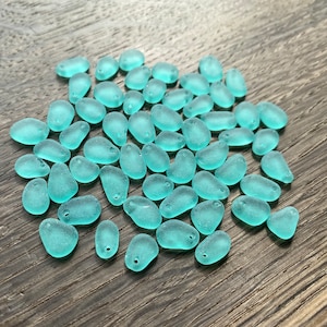 May include: A collection of teal sea glass beads, each with a unique shape and size. The beads are smooth and polished, with a slightly frosted finish.