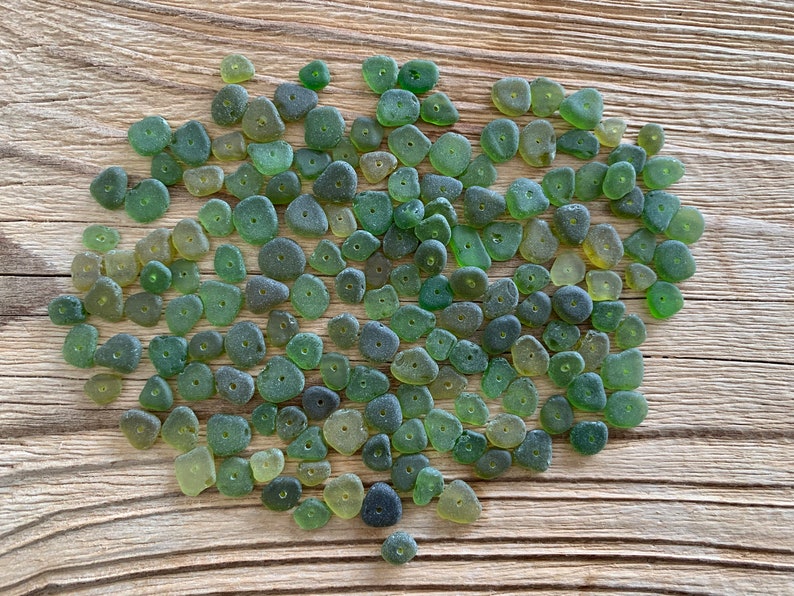 Puede incluir: Un conjunto de cuentas de vidrio marino verdes, en tonos que van desde el oliva claro hasta el verde bosque oscuro, sobre una superficie de madera envejecida. Cada cuenta es ovalada con un peque&ntilde;o agujero, ideal para joyer&iacute;a o manualidades.