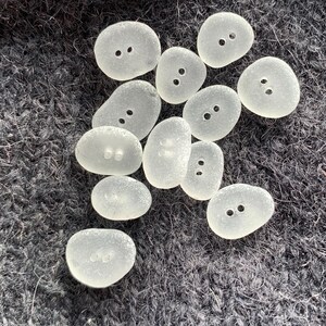Puede incluir: Una colecci&oacute;n de botones de vidrio de mar lisos y blancos, con dos agujeros cada uno. Los botones son de varios tama&ntilde;os y formas, y est&aacute;n dispuestos sobre una superficie gris oscuro y difusa.