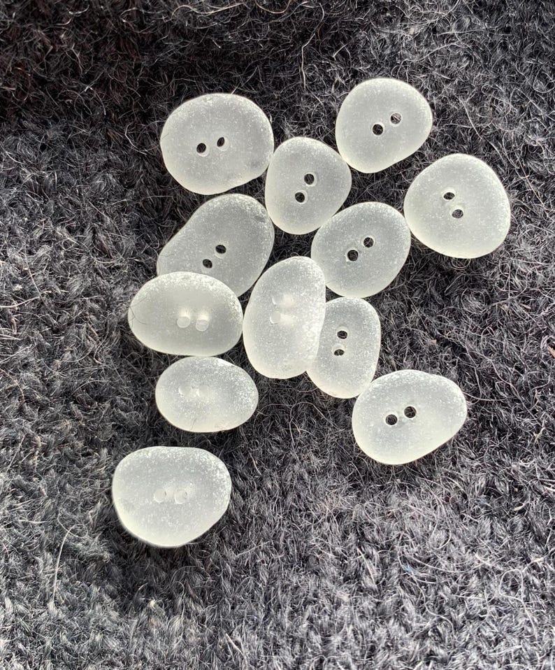Puede incluir: Una colecci&oacute;n de botones de vidrio de mar lisos y blancos, con dos agujeros cada uno. Los botones son de varios tama&ntilde;os y formas, y est&aacute;n dispuestos sobre una superficie gris oscuro y difusa.