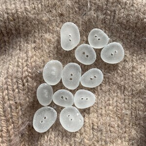 Puede incluir: Una colecci&oacute;n de 11 botones de vidrio lisos y blancos, con dos agujeros cada uno. Los botones est&aacute;n dispuestos en un c&iacute;rculo suelto sobre una superficie marr&oacute;n y peluda.