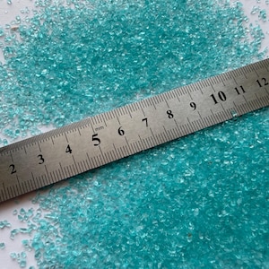 May include: A pile of small, translucent turquoise glass granules scattered across a surface. A stainless steel ruler is placed diagonally across the granules, with the scale clearly showing measurements in centimeters up to 14 cm in length.