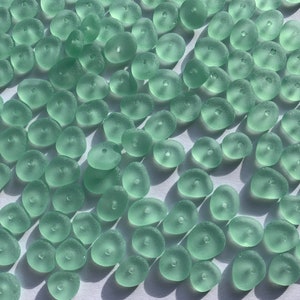 Op de afbeelding: Een verzameling lichtgroene zeeglas kralen met een gat in het midden van elke kraal. De kralen zijn glad en hebben een matte afwerking.