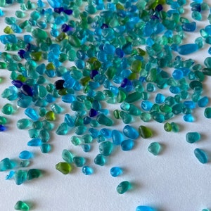 May include: A collection of small, colorful sea glass pieces in shades of blue, green, and turquoise. The sea glass is scattered on a white surface.