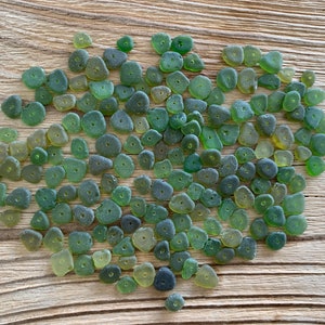 Puede incluir: Una colecci&oacute;n de peque&ntilde;as cuentas de vidrio verde mar con un agujero en el centro. Las cuentas var&iacute;an en forma y tama&ntilde;o, y est&aacute;n dispersas sobre una superficie de madera marr&oacute;n claro envejecida. Las cuentas tienen un aspecto esmerilado.