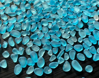 May include: A large collection of small, smooth, frosted sea glass pieces in various shades of bright cyan and light blue. Each piece features rounded edges and a translucent, matte texture, scattered across the frame in an irregular pattern.