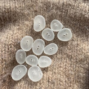 Puede incluir: Una colecci&oacute;n de 11 botones de vidrio de mar liso y blanco. Los botones son de varias formas y tama&ntilde;os, con dos agujeros cada uno. Los botones est&aacute;n dispuestos sobre una superficie de punto marr&oacute;n.