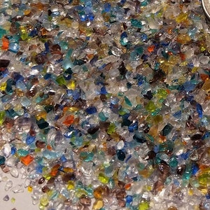 Puede incluir: Primer plano de un montón de coloridos trozos de vidrio triturado. Los fragmentos de vidrio son de varios colores, incluyendo azul, verde, naranja, amarillo y transparente. Las piezas tienen forma irregular y varían en tamaño, creando una apariencia texturizada.