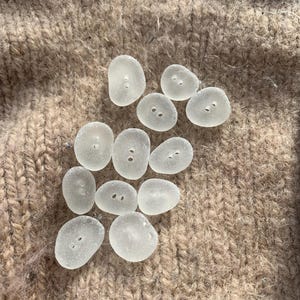 Puede incluir: Una colecci&oacute;n de diez botones de vidrio de mar liso, blanco y transparente, con dos agujeros cada uno. Los botones est&aacute;n dispuestos sobre una tela de punto marr&oacute;n.