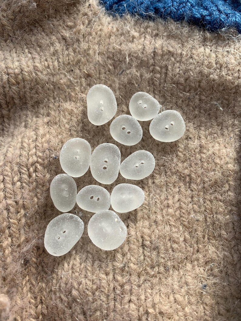 Puede incluir: Una colecci&oacute;n de diez botones de vidrio de mar liso, blanco y transparente, con dos agujeros cada uno. Los botones est&aacute;n dispuestos sobre una tela de punto marr&oacute;n.