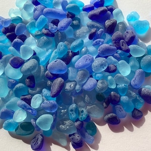 May include: A collection of small, smooth, sea glass pieces in shades of blue, teal, and purple. The glass pieces are scattered on a white surface.