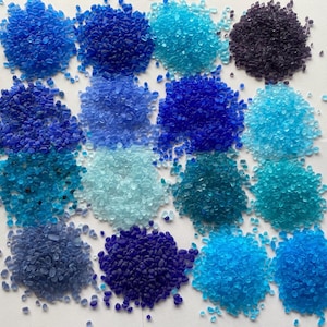 Puede incluir: Vista cenital de una colección de pequeños trozos de vidrio de forma irregular en varios tonos de azul. Los colores varían del turquesa claro al índigo oscuro, dispuestos en montones separados. Las piezas parecen ser suministros decorativos o artesanales.