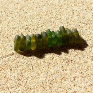 Puede incluir: Una hilera de cuentas de vidrio marino verdes de diferentes tonos, dispuestas sobre arena beige clara. Las cuentas son lisas y redondeadas. La luz del sol proyecta una sombra, resaltando la textura de la arena y las cuentas.