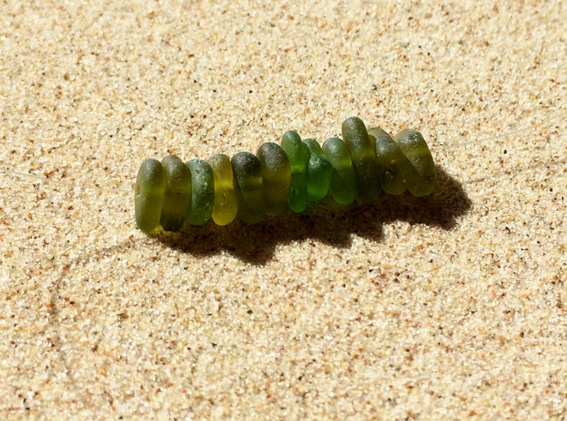 Puede incluir: Una hilera de cuentas de vidrio marino verdes de diferentes tonos, dispuestas sobre arena beige clara. Las cuentas son lisas y redondeadas. La luz del sol proyecta una sombra, resaltando la textura de la arena y las cuentas.
