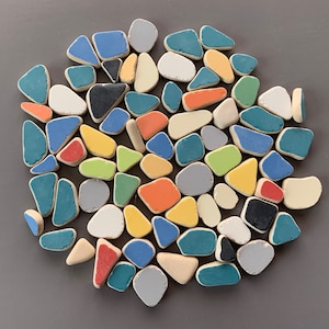 以下が含まれることがあります： 青、グレー、オレンジ、黄色、緑など、さまざまな色で塗装された、不規則な形のセラミック片のコレクション。これらのピースは、灰色の背景に対して円形のパターンに配置され、モザイク効果を生み出しています。