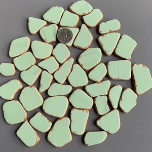 May include: A collection of irregularly shaped, light green ceramic pieces with a quarter coin for size reference. The pieces have a distressed, aged appearance with exposed brown edges, arranged on a gray surface. The ceramic pieces are likely for crafting or decorative purposes.