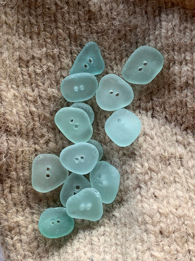 K&ouml;nnte beinhalten: Eine Sammlung von hellblauen, glatten, polierten Meeresglas-Kn&ouml;pfen mit jeweils zwei L&ouml;chern. Die Kn&ouml;pfe haben eine unregelm&auml;&szlig;ige Form.