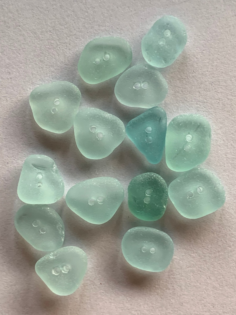 K&ouml;nnte beinhalten: Eine Sammlung von 15 glatten, hellblauen Meerglass-Kn&ouml;pfen mit jeweils zwei L&ouml;chern. Die Kn&ouml;pfe sind in verschiedenen Formen und Gr&ouml;&szlig;en, einige sind runder und andere dreieckiger.