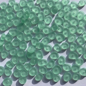Op de afbeelding: Een grote groep lichtgroene zeeglas kralen. De kralen zijn glad en hebben een matte afwerking. Ze zijn allemaal in verschillende vormen en maten.