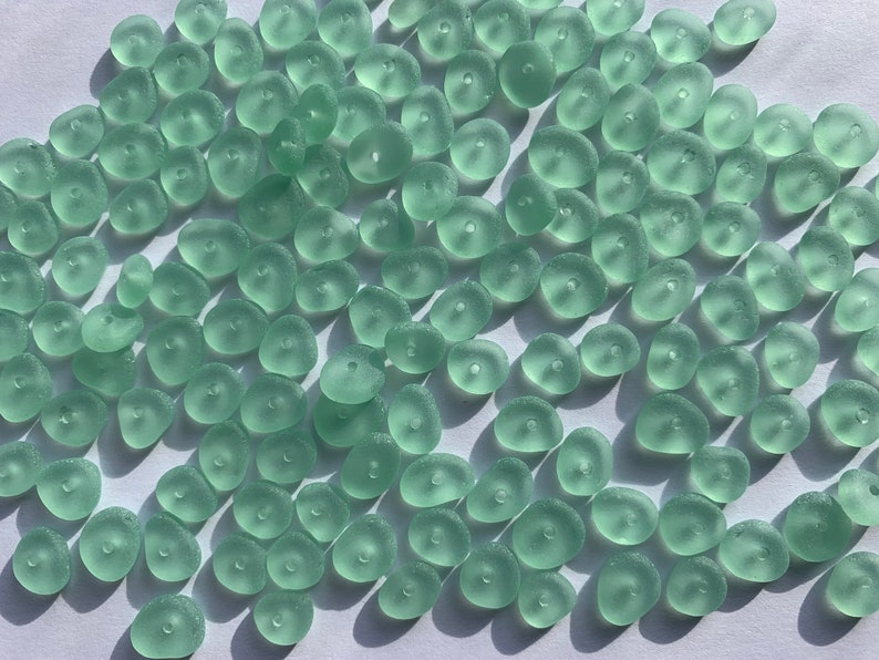 Op de afbeelding: Een grote groep lichtgroene zeeglas kralen. De kralen zijn glad en hebben een matte afwerking. Ze zijn allemaal in verschillende vormen en maten.