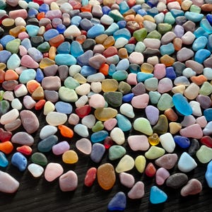 May include: A collection of colorful, smooth, and polished sea glass stones in various shapes and sizes. The sea glass is arranged on a dark surface.