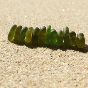 Puede incluir: Un collar de cuentas de vidrio marino en varios tonos de verde, ensartadas en un cord&oacute;n fino y transparente. Las cuentas son oblongas y lisas, descansando sobre un lecho de arena clara. La luz natural crea reflejos y sombras.