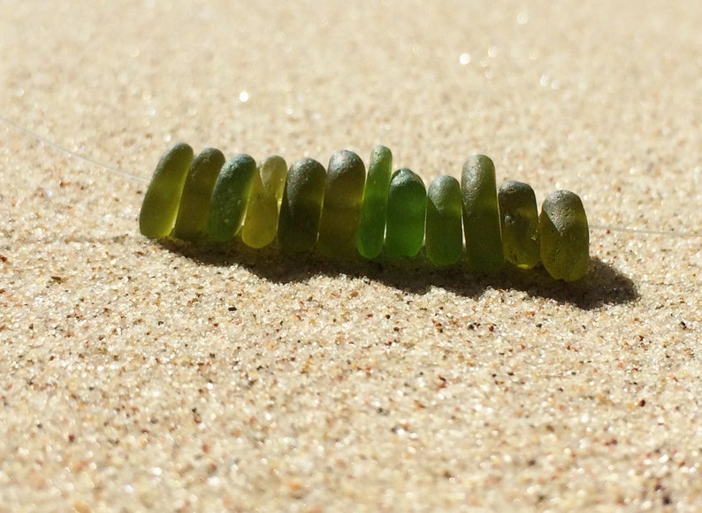Puede incluir: Un collar de cuentas de vidrio marino en varios tonos de verde, ensartadas en un cord&oacute;n fino y transparente. Las cuentas son oblongas y lisas, descansando sobre un lecho de arena clara. La luz natural crea reflejos y sombras.
