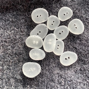 Puede incluir: Una colecci&oacute;n de botones de vidrio de mar liso y blanco, con dos agujeros cada uno. Los botones est&aacute;n dispuestos en un grupo sobre una superficie gris oscuro y difusa.