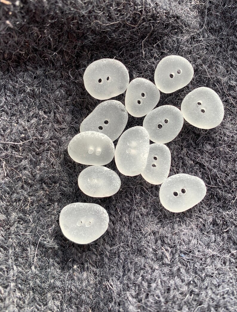 Puede incluir: Una colecci&oacute;n de botones de vidrio de mar liso y blanco, con dos agujeros cada uno. Los botones est&aacute;n dispuestos en un grupo sobre una superficie gris oscuro y difusa.